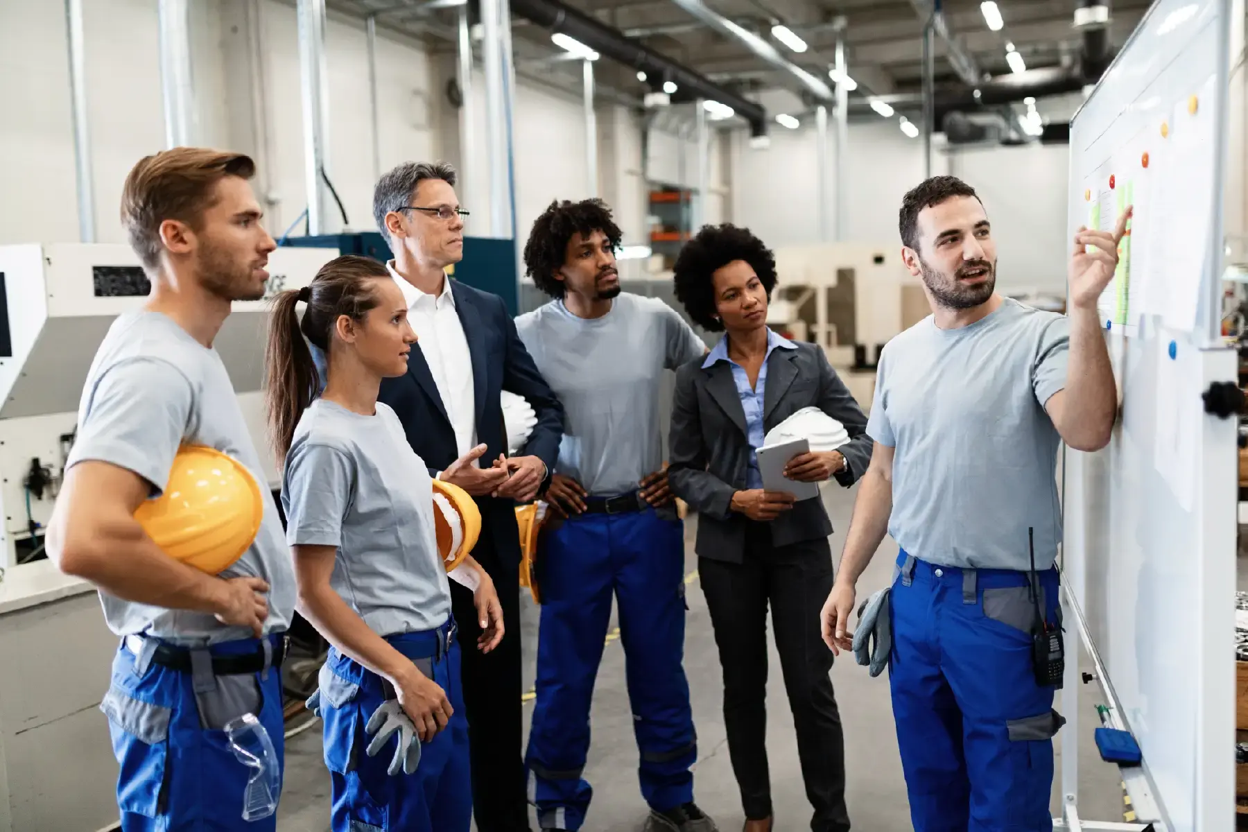 A diverse group of engineers and manufacturing staff align around a shared visual plan — reflecting the internal culture and communication challenges within a global production environment
